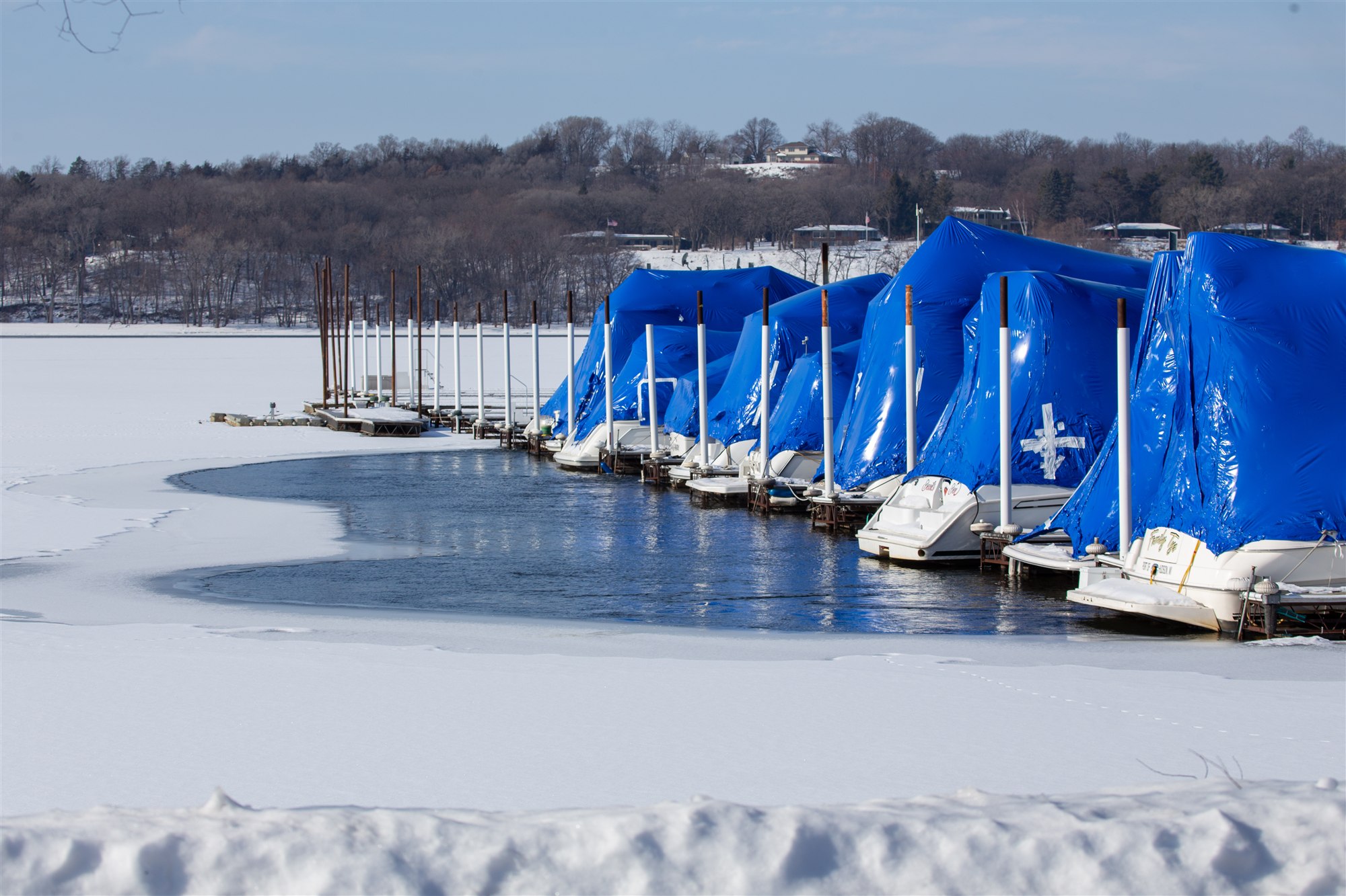 Open water beside a row of blue-covered boats, surrounded by a frozen, snow-covered marina
